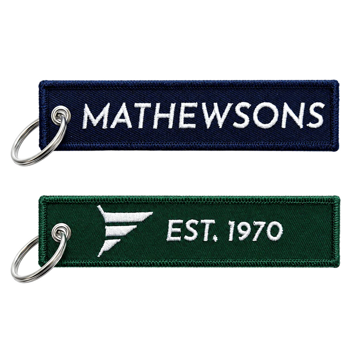 Two embroidered rectangular keychains on a white background. The top one is navy blue with white text reading 'MATHEWSONS' and a silver split ring.